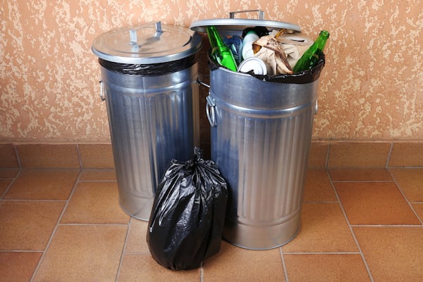 Two aluminum garbage cans full of garbage, including bottles and cloth. Black plastic garbage bag sitting between the two cans. 