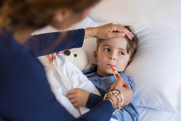 Sick child in bed. Woman feeling his head and taking his temperature with an oral thermometer. Teddy bear tucked into bed next to child.